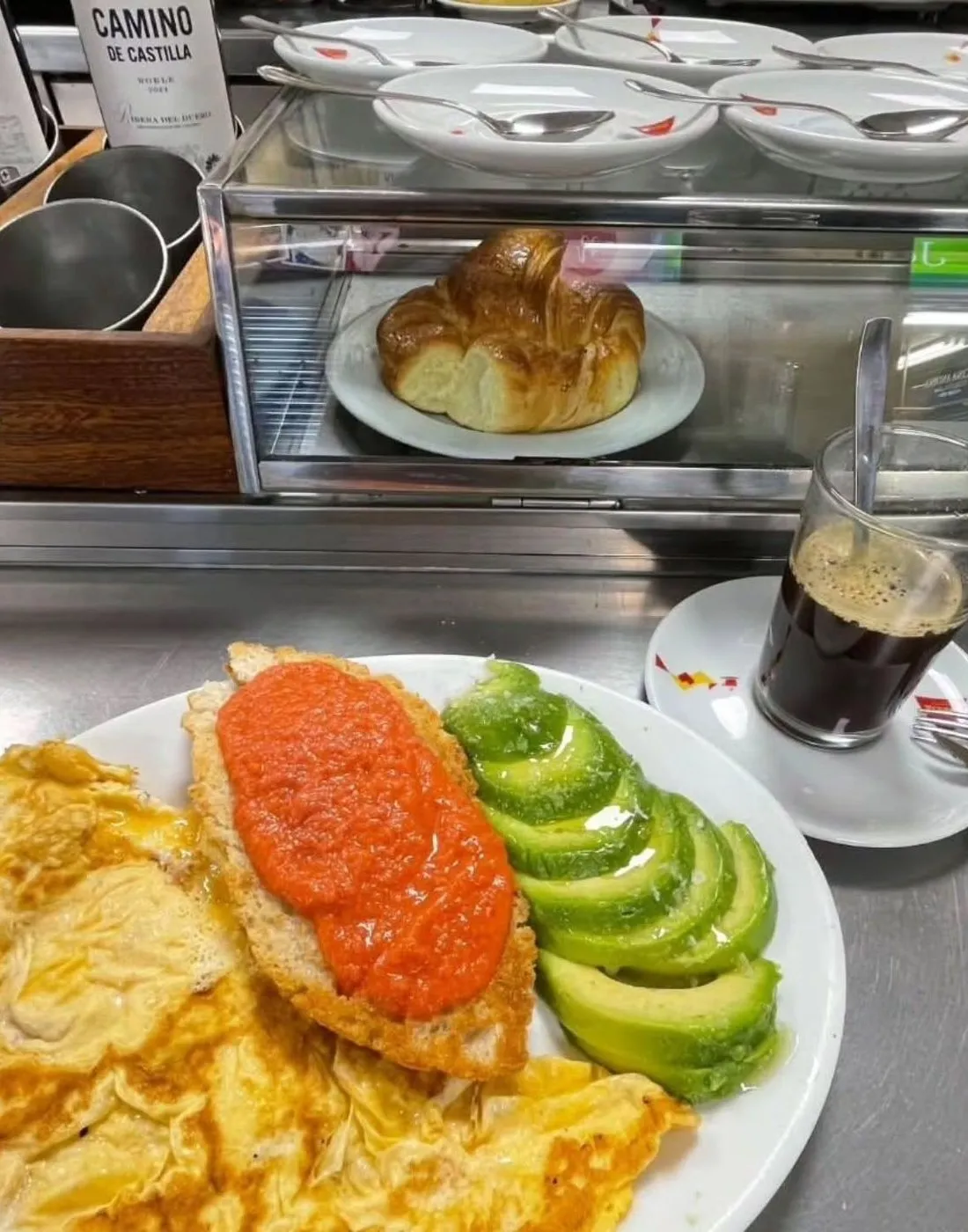 Huevos revueltos, tostada con tomate y aguacate en un plato, con un café y un cruasán detrás